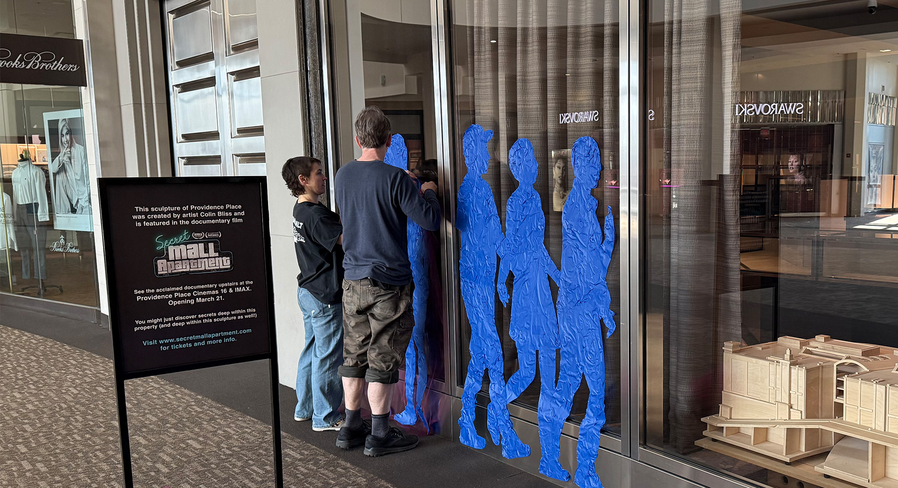 Smith, left, and Townsend install tape-art silhouettes at Providence Place. Beside them is a wooden scale model of the mall c